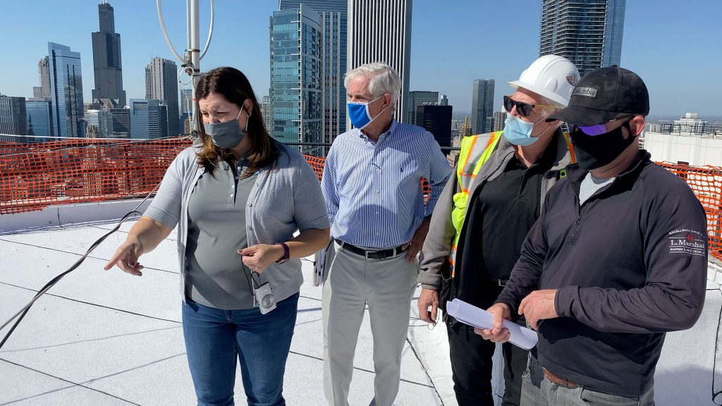 Sonia Surlej of Walker Consultants stands on the roof of a 54-story building and examines the progress of a roof replacement project with representatives from the manufacturer and contractor. Nearby skyscrapers and the Chicago skyline are visible in the background