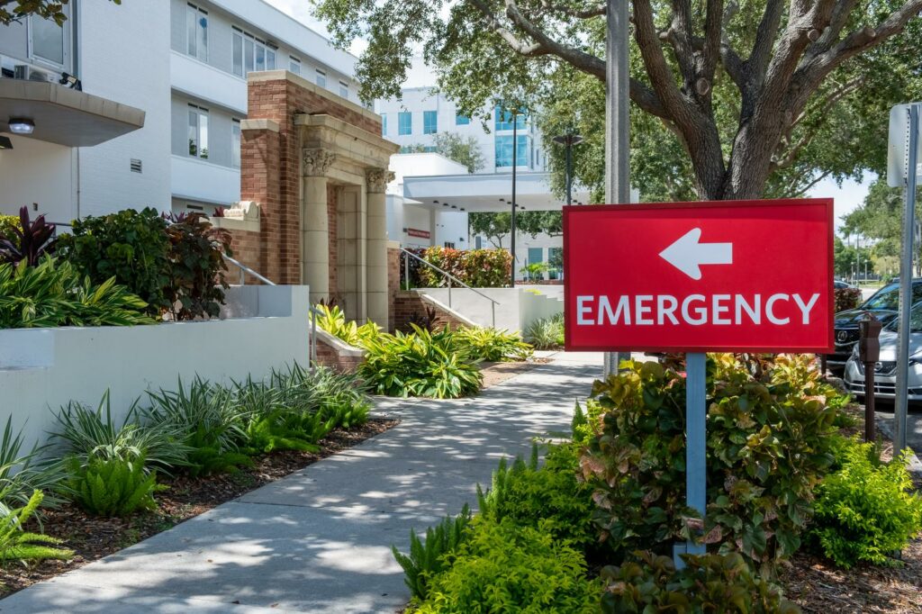 A pleasant sidewalk under a canopy of trees outside a hospital, with a sign pointing to the Emergency department