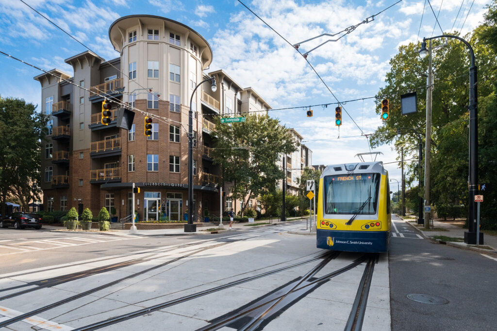 A light rail train in the street passes through an intersection in front of a five-story multifamily housing structure on a sunny day in Charlotte, NC