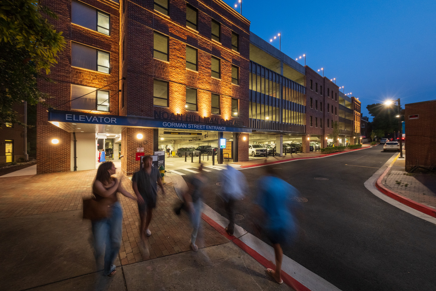 People walk on the sidewalk in front of the Hillman garage on a summer evening
